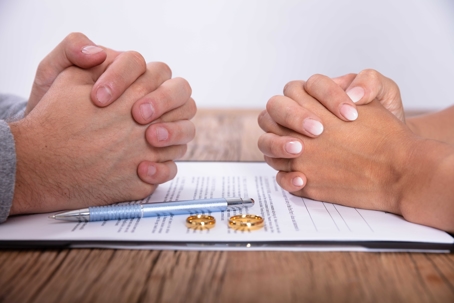 two people fold their hands on either side of a divorce agreement with two gold wedding bands and a pen sitting on top