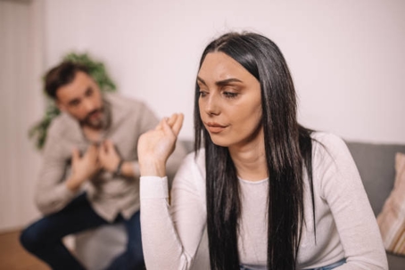 A woman sits on a couch looking away with an upset expression while a man in the background gestures toward her as if trying to talk.