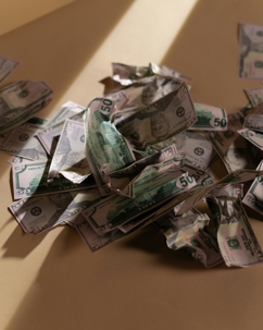 A pile of crumpled U.S. dollar bills scattered on a beige surface with light and shadow across them.