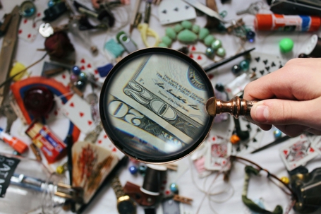 Hand holding a magnifying glass over two twenty-dollar bills, enlarging the details, with a cluttered background of playing cards, jewelry, tools, and small objects scattered on a table.
