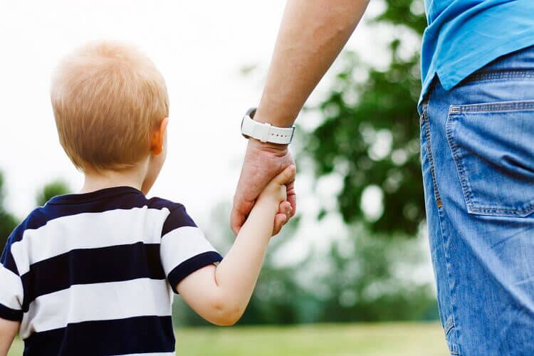 A parent with sole custody walks through a park while holding their small child's hand.